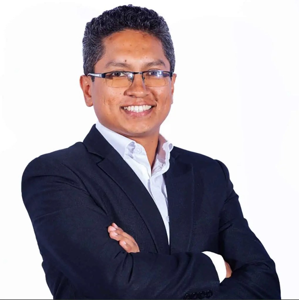 Hombre sonriendo con gafas, traje negro y camiseta blanca, con los brazos cruzados sobre el pecho, frente a un fondo blanco.