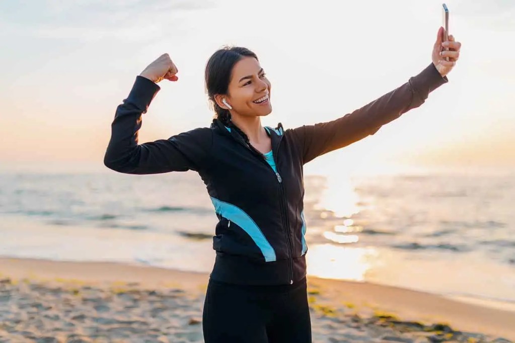 Mujer sonriendo mientras toma un selfie en la playa al atardecer, levantando el brazo en señal de triunfo.