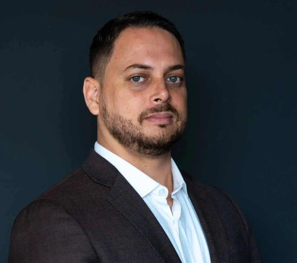 Retrato de un hombre con barba, vestido con un saco oscuro y camisa blanca, sonriendo, con fondo gris oscuro.