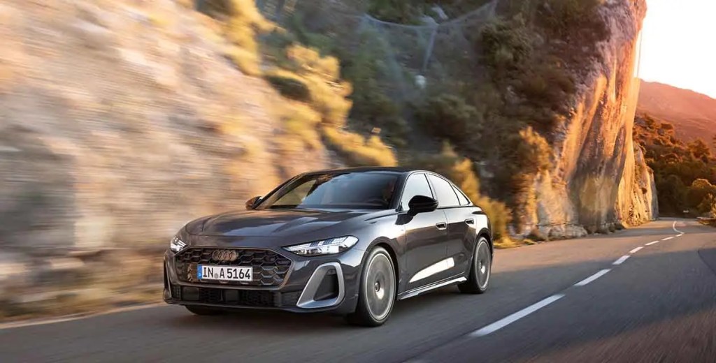 Coche deportivo gris conduciendo por una carretera junto a un acantilado iluminado por la luz del atardecer.