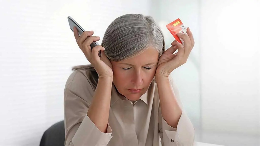 Mujer de mediana edad con cabello canoso, sosteniendo un teléfono móvil y un paquete de medicación, luciendo estresada y con la cabeza inclinada hacia abajo.
