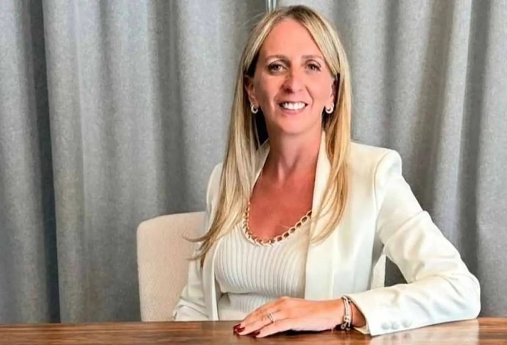 Mujer sonriente sentada en una mesa, vistiendo un blazer blanco y una blusa de rayas, con cortinas grises de fondo.