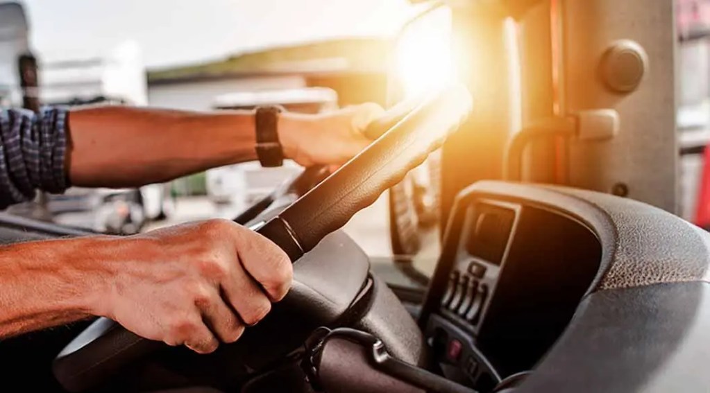 Manos sujetando el volante de un camión en el interior del vehículo, con luz del sol iluminando el escenario.