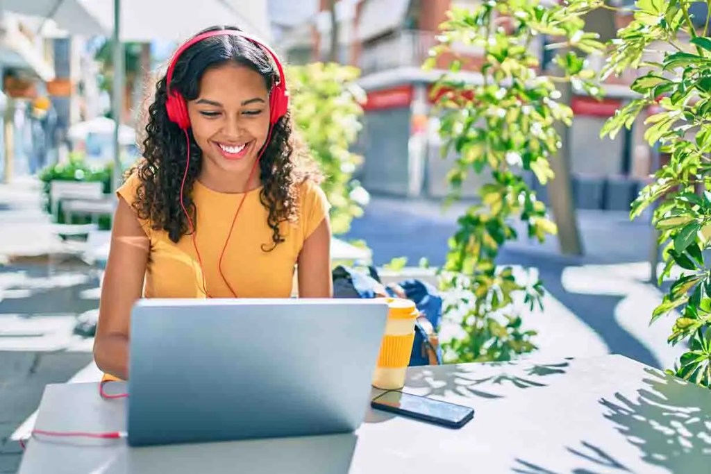 Mujer sonriente con auriculares rojos trabajando en una computadora portátil en una terraza, rodeada de plantas.