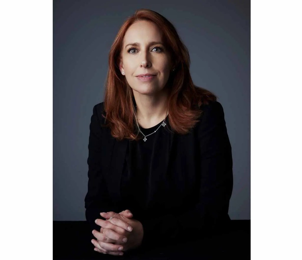 Retrato de mujer con cabello castaño claro, vestida con una blusa negra. Ella está mirando a la cámara con una expresión seria y manos entrelazadas sobre una mesa.