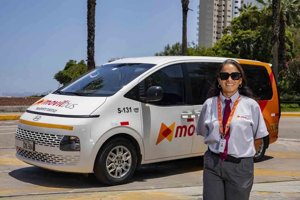 Mujer sonriente vestida con uniforme de conductor de transporte público, de pie frente a una van de movitBus en un entorno urbano.