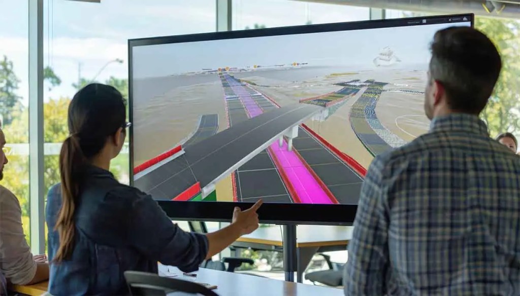 Dos personas observan una pantalla grande que muestra un modelo digital de un proyecto de infraestructura, con caminos y zonas de diferentes colores.