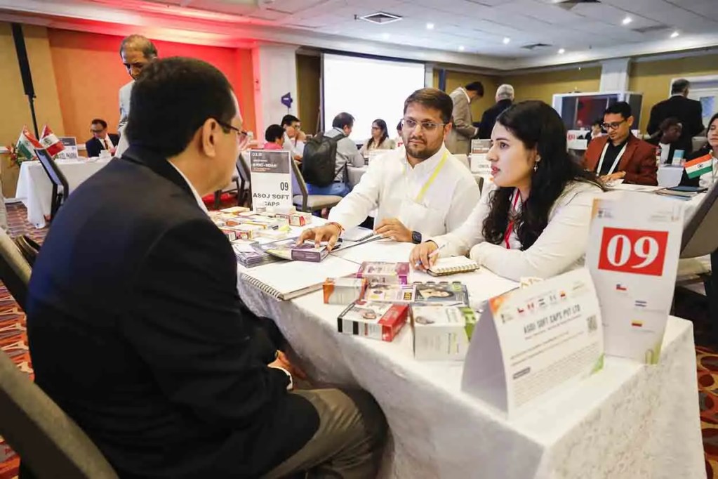 Tres personas conversando en una mesa, discutiendo productos en un evento empresarial. Hay muestras de productos sobre la mesa y banderas visibles en el fondo.