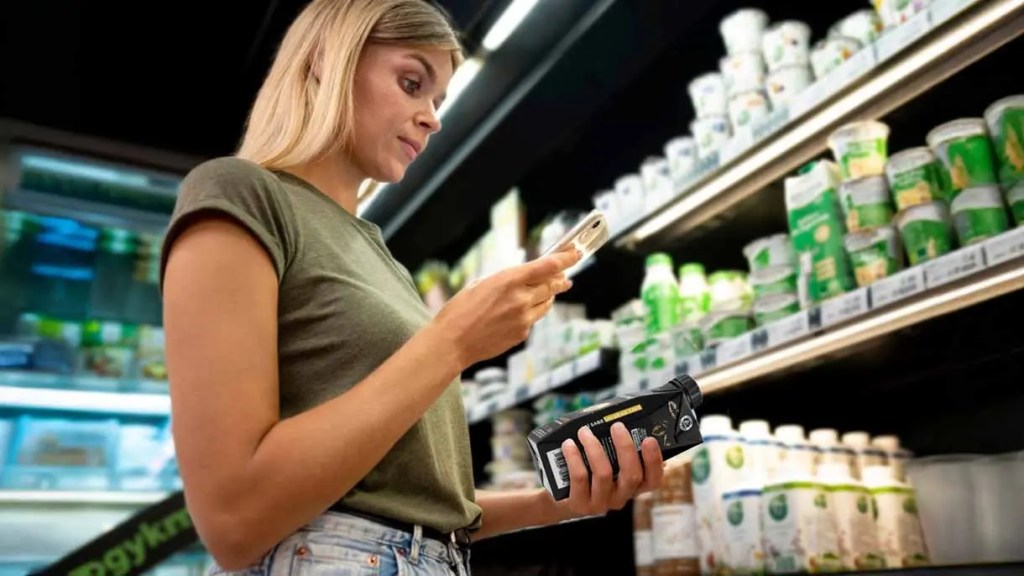 Mujer con cabello rubio revisando su teléfono mientras sostiene un producto en un supermercado, rodeada de estantes con yogures.