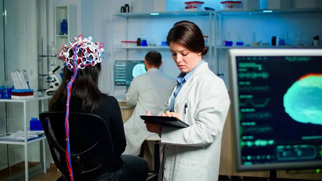 Mujer en bata de laboratorio observando una tablet mientras otra persona con un dispositivo en la cabeza está de espaldas en un laboratorio.