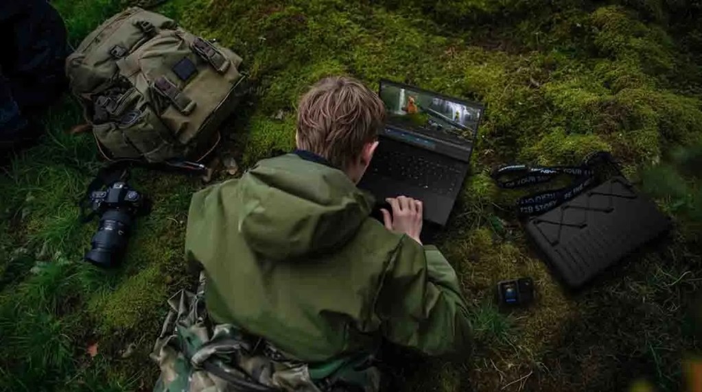 Una persona con abrigo verde trabajando en una laptop sobre musgo, junto a una mochila y una cámara en un entorno natural.