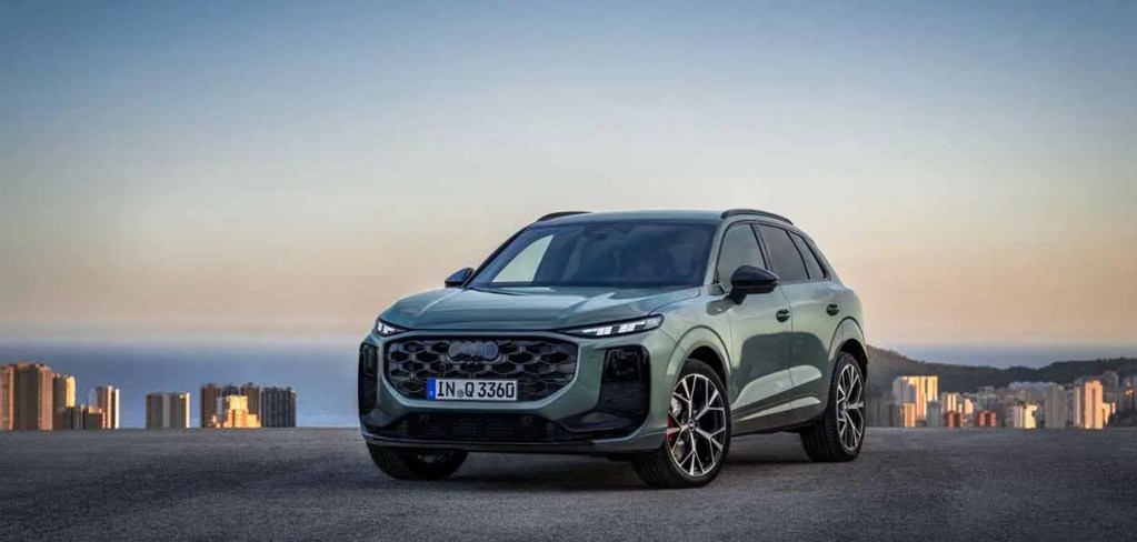 Un SUV de color verde, modelo Audi, estacionado en una carretera con vista a una ciudad costera al atardecer.