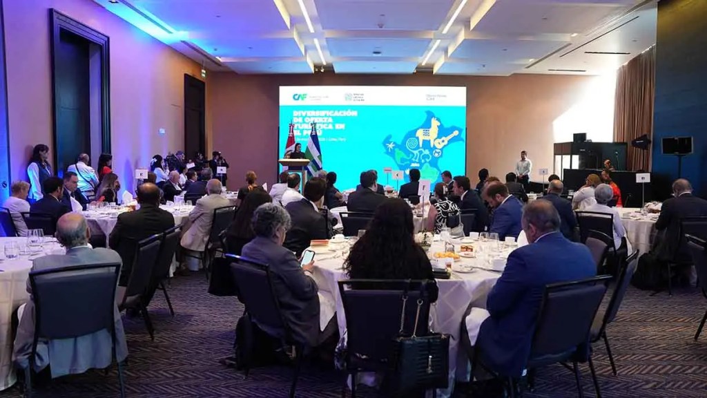 Evento con diversas mesas y asistentes disfrutando de un almuerzo, mientras un orador presenta en un escenario con una pantalla detrás que muestra el tema 'Diversificación de Origen Turística en México'.