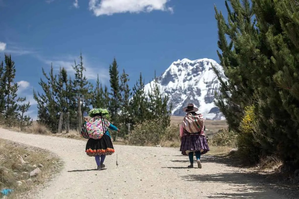 Dos mujeres indígenas caminan por un camino de tierra con un paisaje montañoso nevado al fondo.