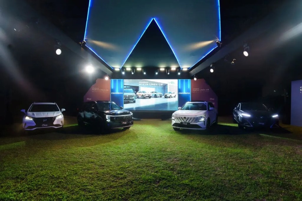 Exhibición nocturna de cuatro automóviles en un evento, con un fondo iluminado y pantallas que muestran información sobre los vehículos.