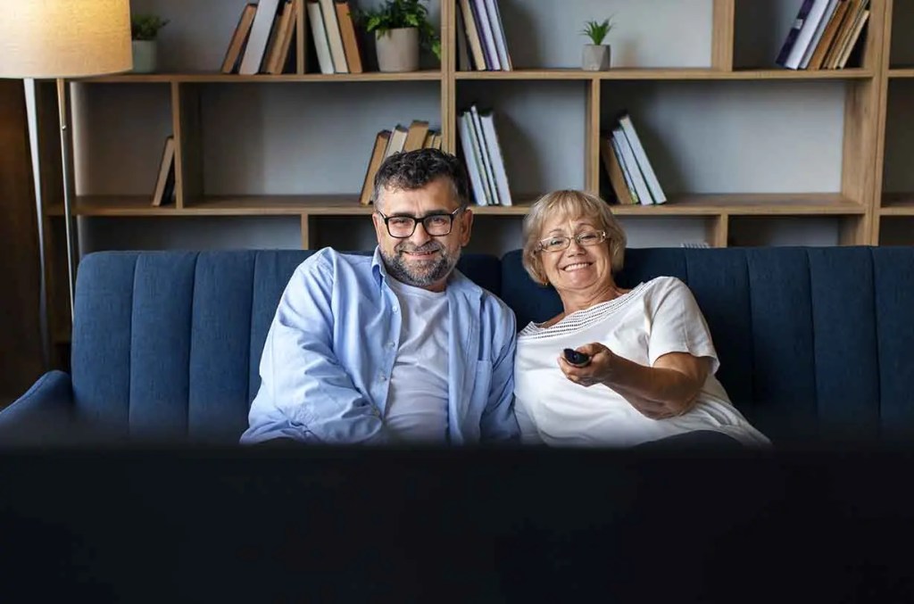 Pareja mayor sentada en un sofá viendo televisión y sonriendo.