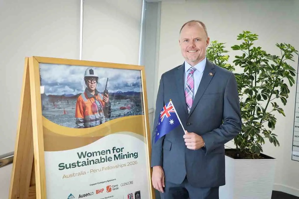 Hombre de pie con una bandera de Australia junto a un cartel que dice 'Women for Sustainable Mining' en un entorno de oficina con plantas.