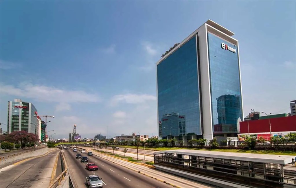 Vista de una avenida con edificios modernos y un rascacielos de vidrio en el fondo, bajo un cielo despejado.