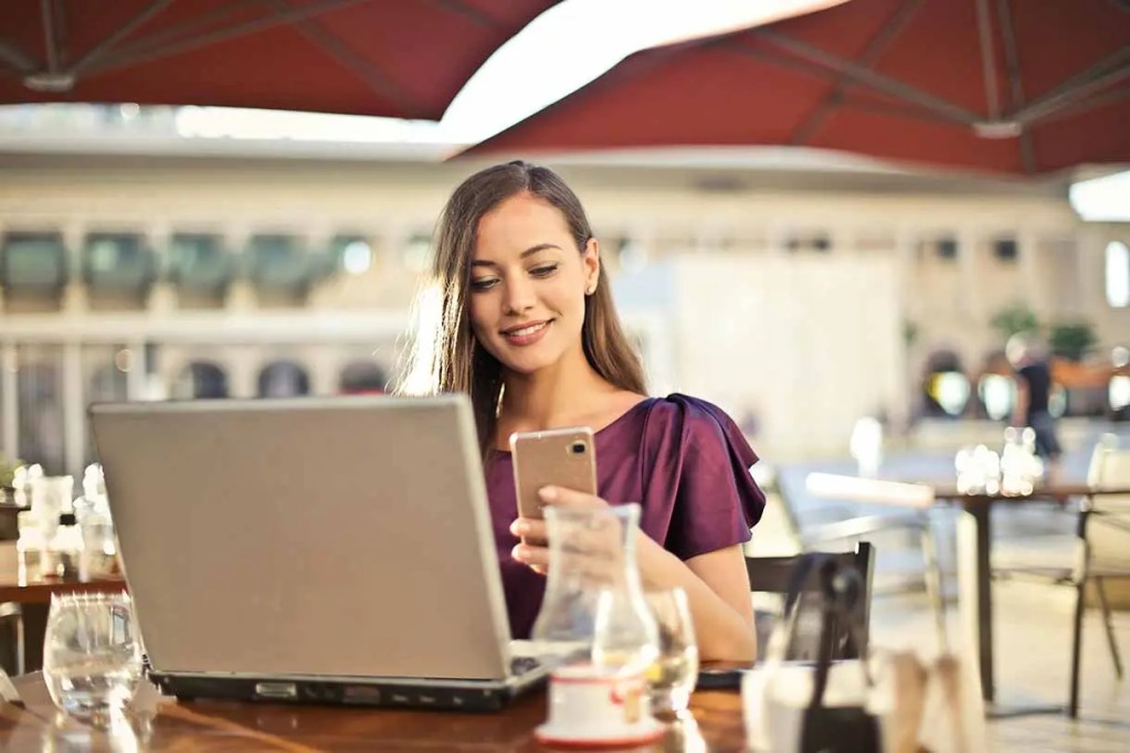 Mujer sonriente sentada en una mesa de café, usando un teléfono móvil mientras mira una computadora portátil.