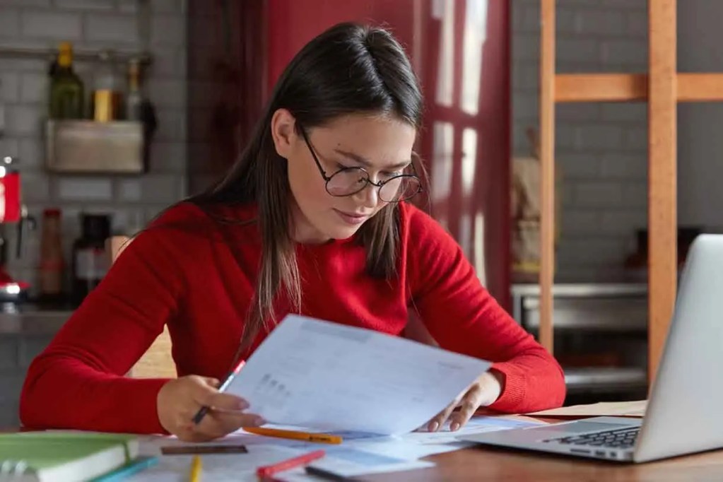 Una mujer con gafas está revisando documentos en una mesa de trabajo, frente a una computadora portátil en un ambiente de cocina.