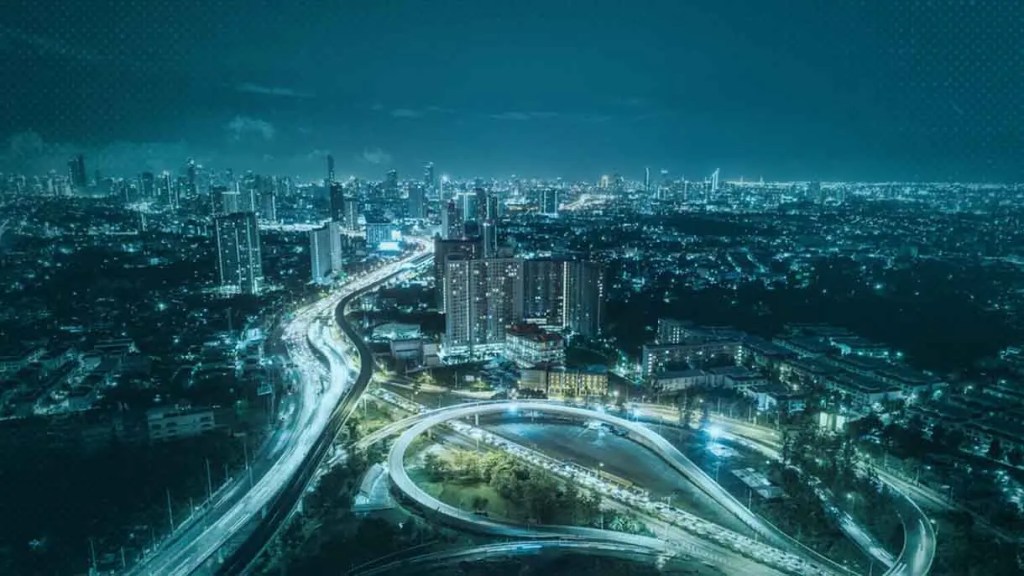 Vista nocturna de una ciudad iluminada, con carreteras y edificios en primer plano. Se observan luces de tráfico y una atmósfera urbana.