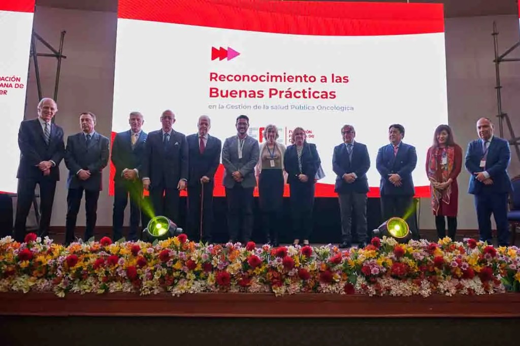 Grupo de personas en un evento de reconocimiento a las Buenas Prácticas en la Gestión de la Salud Pública Oncológica, con un fondo de flores coloridas y una pantalla detrás mostrando el título del evento.
