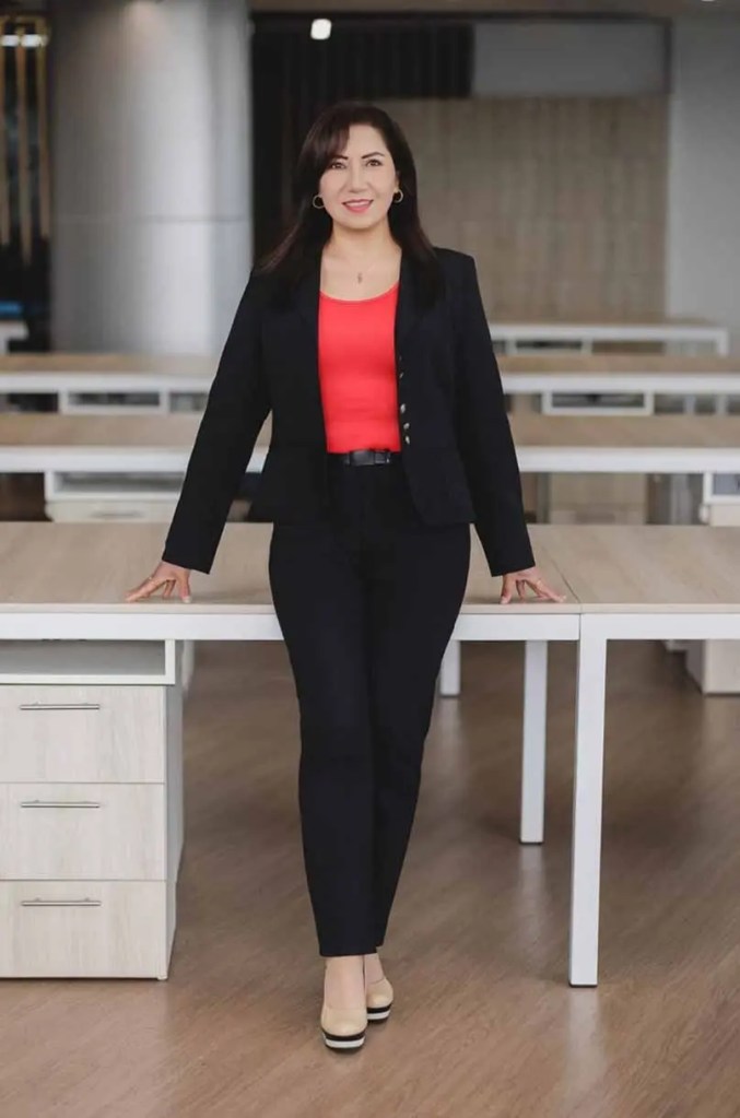 Una mujer de pie frente a un escritorio, vistiendo un traje negro y una blusa roja, sonriendo mientras apoya las manos en la superficie del escritorio.