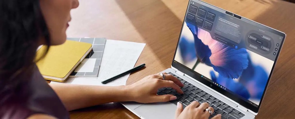 Persona utilizando un laptop con pantalla de fondo de una flor, sobre una mesa de madera con una libreta amarilla y documentos.