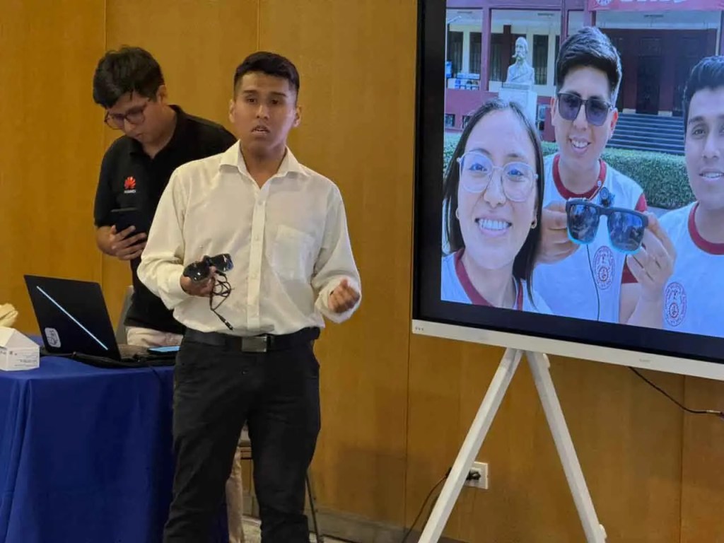Un joven presenta un proyecto mientras sostiene unos lentes, con una pantalla detrás que muestra una selfie de él y otros compañeros en un evento educativo.