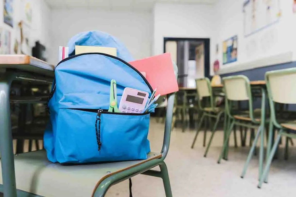 Una mochila azul con útiles escolares se encuentra sobre una silla en un aula vacía.
