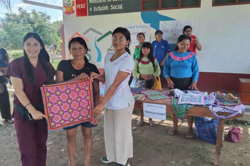 Tres mujeres sostienen una tela artesanal colorida en un evento comunitario en Perú, mientras que otras personas participan en el fondo. Se observa un cartel del Ministerio de Desarrollo e Inclusión Social.