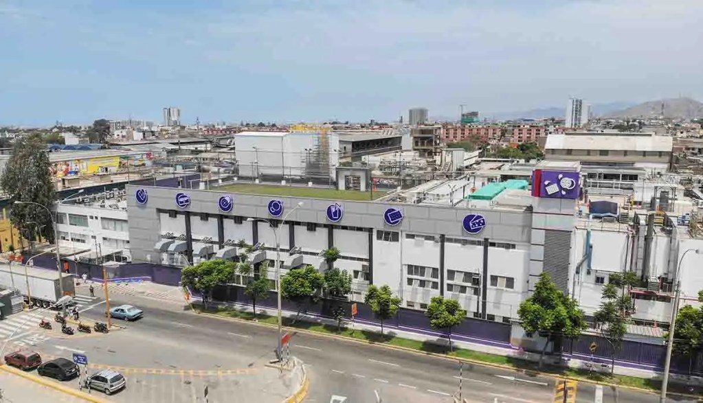 Vista aérea de un edificio industrial moderno con logotipos en la fachada, ubicado en un área urbana rodeada de otras estructuras y vegetación.