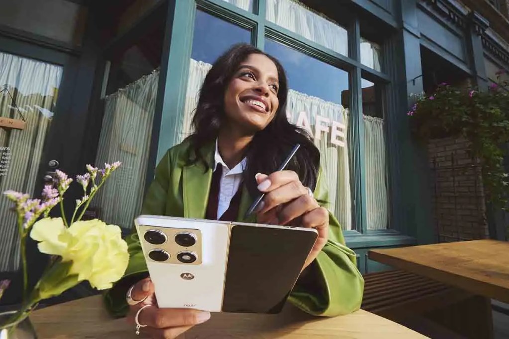 Mujer sonriendo mientras sostiene un teléfono móvil y un bolígrafo en una cafetería al aire libre, con flores en la mesa.