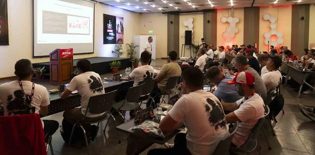 Una sala de conferencias con varias personas sentadas, escuchando a un ponente frente a una pantalla. Las personas llevan camisetas de color claro y hay plantas en las mesas.