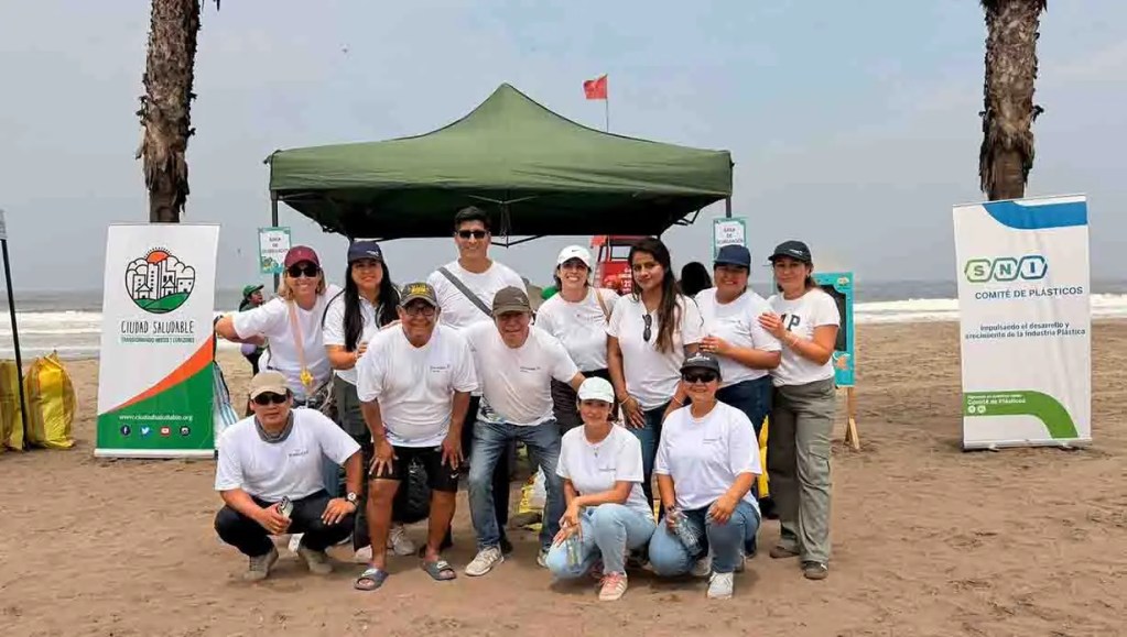 Grupo de personas en la playa frente a una carpa verde, participando en una actividad comunitaria, con banderas y carteles relacionados con la limpieza y el medio ambiente.