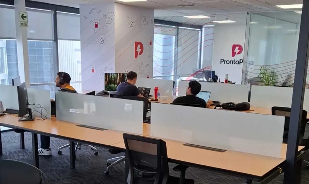 Oficina moderna con trabajadores en escritorios, pantallas de computadora y logo de ProntoP en la pared.