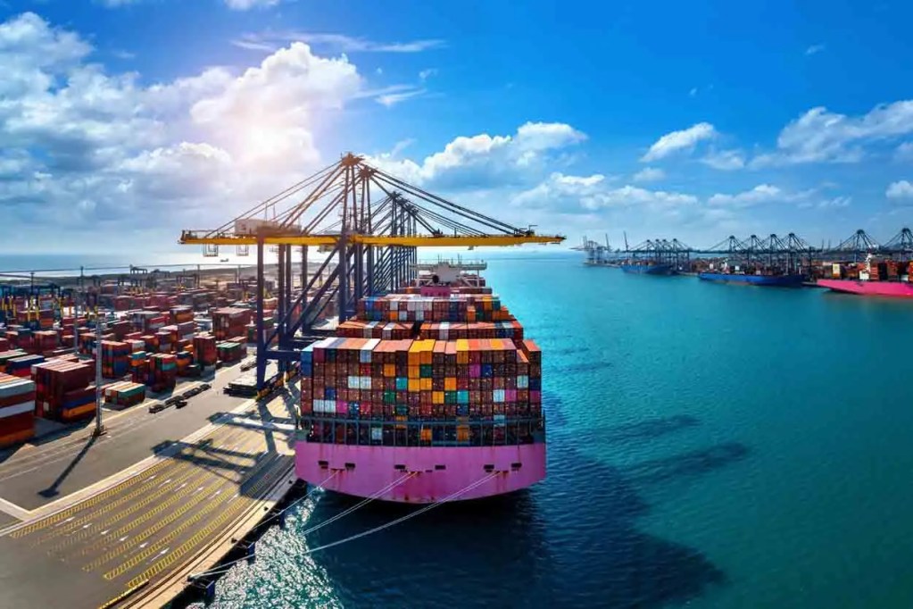 Vista aérea de un puerto con un barco de carga cargado de contenedores coloridos, grúas trabajando y un cielo azul con nubes.