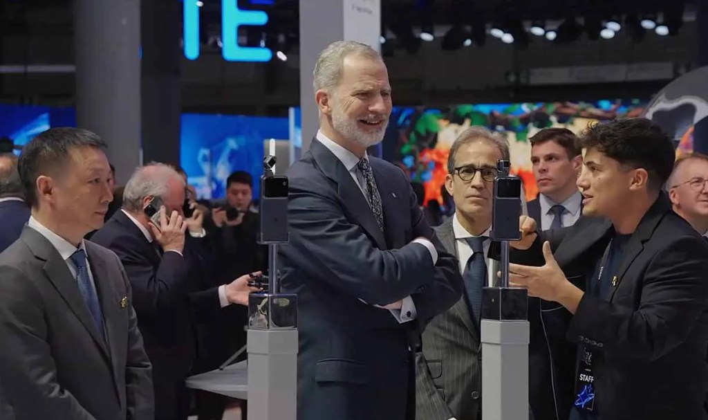 Un grupo de personas en una feria tecnológica, con un hombre mostrando un dispositivo a un grupo que incluye a una figura prominente sonriente.