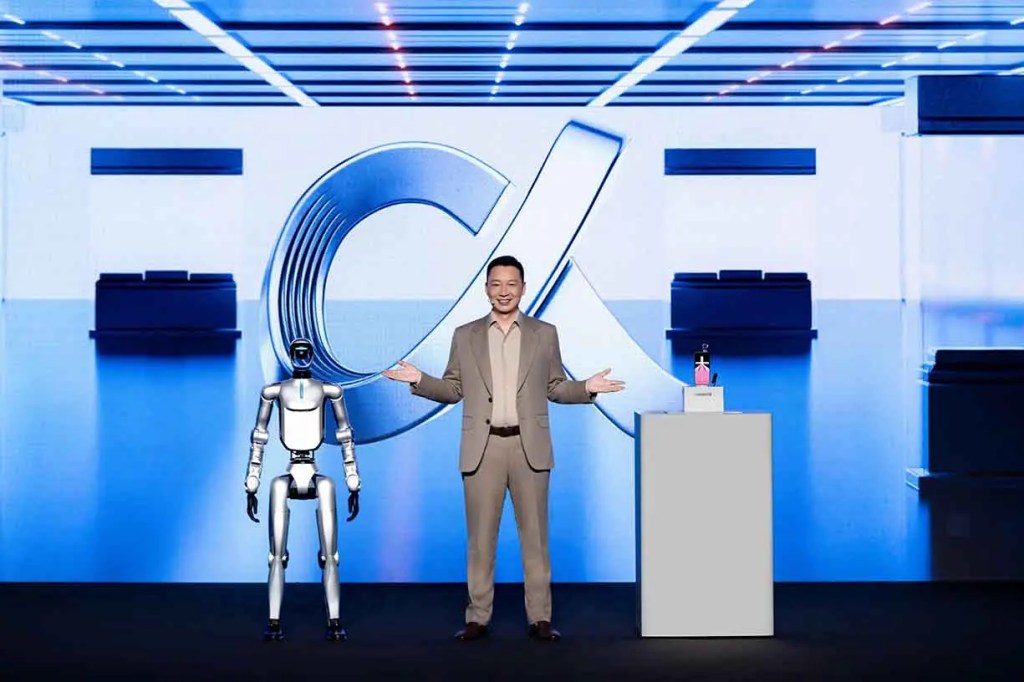 Un hombre sonriente presenta un robot en un escenario tecnológico, con un gran logo de fondo y un dispositivo en una mesa.