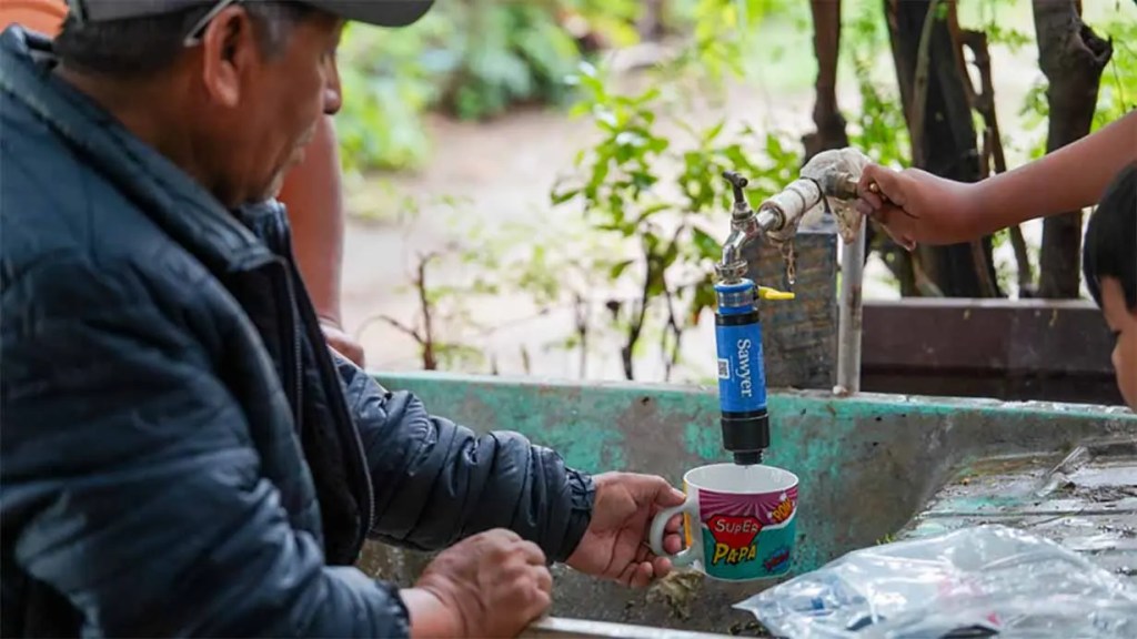 Hombre llenando una taza con agua de un grifo, mientras un niño ayuda con el grifo en un entorno natural.