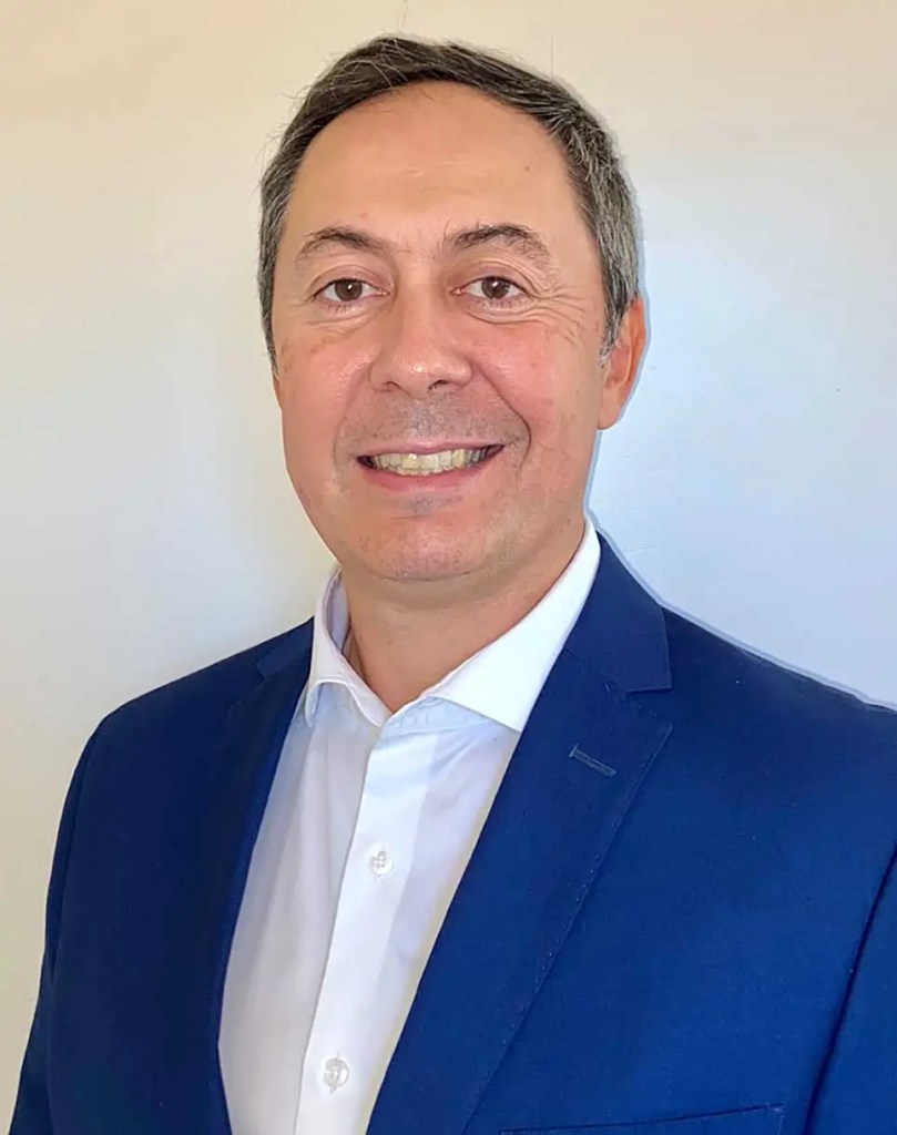 Retrato de un hombre sonriendo, con chaqueta azul y camisa blanca, frente a un fondo claro.
