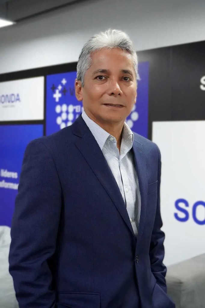 Retrato de un hombre con cabello canoso, vestido con un traje oscuro y camisa de rayas, posando en un ambiente de oficina con fondo azul y blanco.
