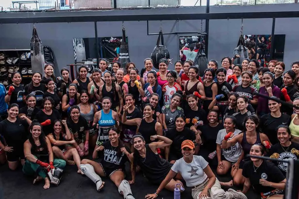Grupo de mujeres en un gimnasio de boxeo, posando juntas con guantes de boxeo, algunas sonriendo y otras haciendo gestos de victoria.
