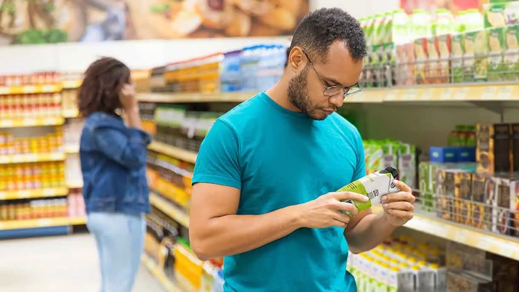 Un hombre con gafas sostiene un envase en un supermercado mientras examina la etiqueta de un producto. En el fondo, una mujer habla por teléfono.