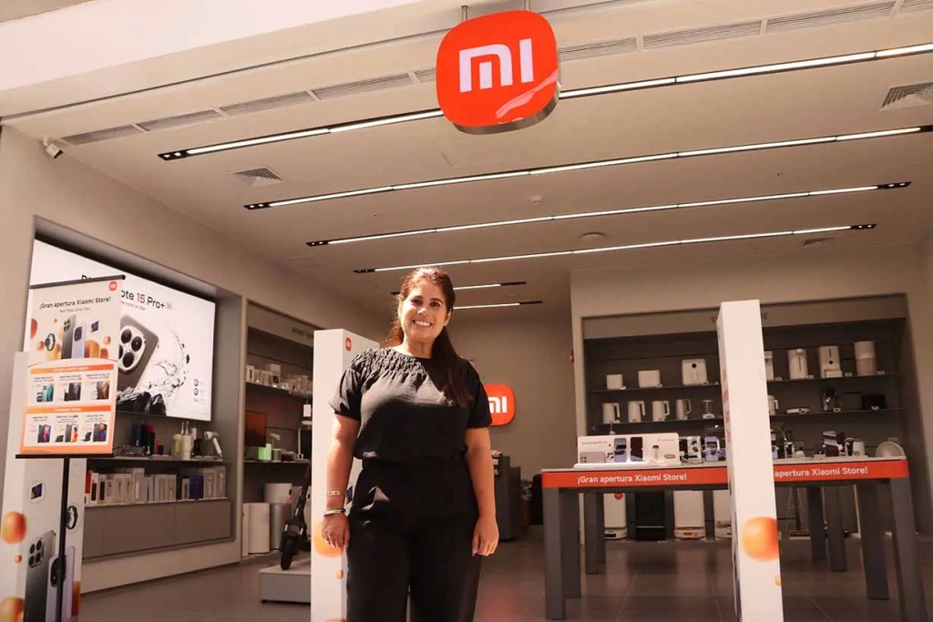Una mujer sonriente de pie frente a una tienda Xiaomi, con productos en exhibición y un letrero rojo con el logotipo de MI en la parte superior.