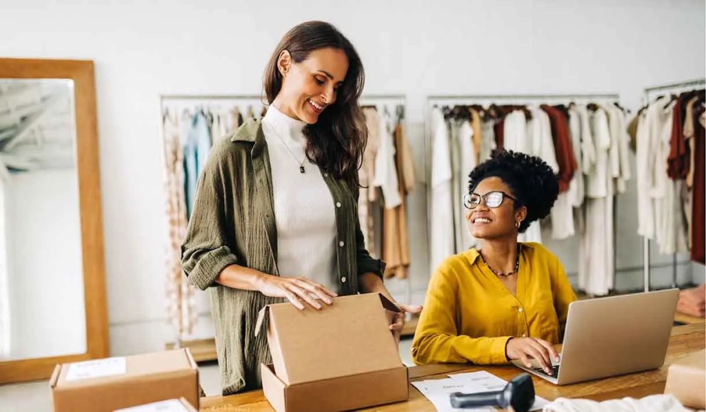 Dos mujeres en una tienda de ropa, una revisando una caja y la otra sonriendo mientras trabaja en una computadora portátil.