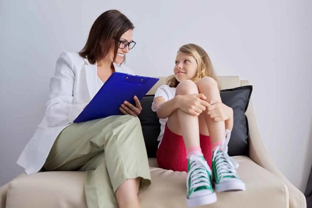 Una terapeuta sonriente, con gafas, está sentada en un sofá junto a una niña, quien se muestra curiosa y cómoda. La terapeuta sostiene una carpeta azul y ambas parecen estar en una conversación amigable.