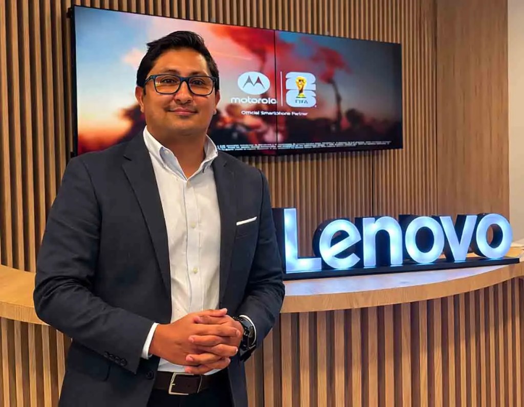 Hombre de pie frente a un letrero de Lenovo en una oficina moderna, con una pantalla que muestra el logo de Motorola y un evento de FIFA detrás de él.