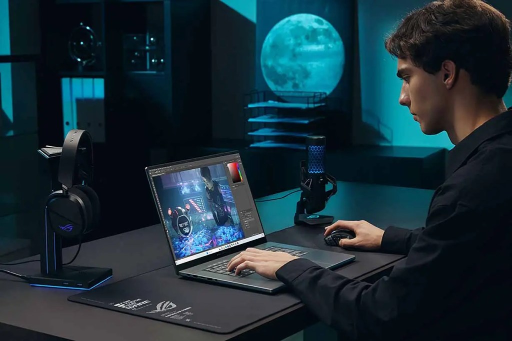 Un joven sentado frente a una computadora portátil, editando imágenes en un entorno de trabajo moderno y iluminado. Hay un micrófono y unos auriculares gaming en la mesa, además de una luna llena visible en el fondo.