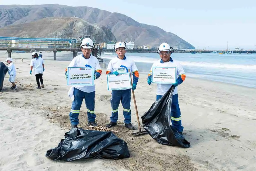 Tres hombres en una playa, vestidos con camisetas y cascos de seguridad, sosteniendo carteles que promueven la protección del medio ambiente. En el fondo, otras personas recogen basura de la playa.